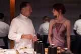 An image shows actress Anya Taylor-Joy in her character of Margot from The Menu. She wears a pink dress and her hair is an in updo. She looks surprised and to her right at Ralph Fiennes, who is in character from the movie. He is in chef’s attire. There are multiple sous-chefs working in the background.