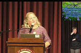 Screenshot from a talk by Robin Wall Kimmerer. She stands behind a podium. In one corner is a mini view of her slides. Below that in another corner is the ASL interpreter.
