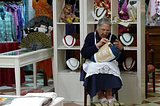 A lacemaker at work. A grandmotherly elder women sits in the center of clothing store and stitches her lace work. She wears a classic white apron with a lace border and is surrounding by many textiles — fans, dresses and also jewelry.