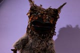An owlbear puppet stands, looking at the camera. They look cheery and fun and about to say something. The arm of the puppeteer is just visible at the bottom of the image.
