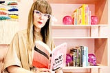 A woman reads the dark satiric wellness influencer novel Self Care by Leigh Stein in a pink room.