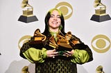 Eilish stands smiling, holding the five grammys she won at the 2020 Grammy Awards