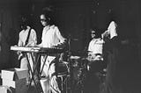 A black and white photo of performers playing musical instrument on stage at the Edinburgh fringe. They are all wearing black sunglasses and white clothes.