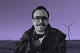 A digitally edited photograph of a man with light skin, a mustache, and glasses, wearing a black leather jacket over a dark shirt. He is smiling slightly and looking directly at the camera. The background features an open rural landscape with wind turbines in the distance. The entire image has been color-edited with a monochromatic purple hue.