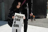 girl holding fight like a girl-printed paper beside woman holding plastic pack