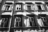 Black and white photograph of the facade of a residential building in Trieste, windows, shutters, shadows — by Russell Davies