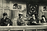 Two Black Panthers sit with two Young Patriots at a table. Free the Panther banners and a confederate flag hang behind them.