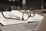 Glasses and a pen on a journal in front of a laptop