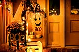 A porch decorated with Halloween decor — ghosts, skeletons, pumpkins, orange and yellow flowers, signs that say “Boo,” and fairy lights.