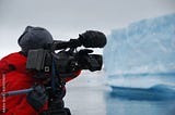 Cameraman wearing red coat, beanie, and gloves points a camera at an iceberg in Antarctica