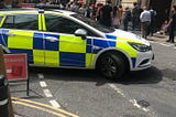 A police car, with a yellow-and-blue Battenberg pattern design, is closing off Clare Street in Bristol city centre to protect Dr Who filming. A crowd watches the filming taking place.