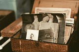The image, washed out, is of a small wooden box perched atop a table. The box is open and inside rest old photos of people long since deceased that tell the story of someone’s life.
