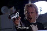A man with bleached blond hair in a tuxedo points a gun in a studio. He shouts, ‘Power to the people who punish bad cinema!’