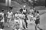 Black and white photo. Kathrine Switzer became the first woman to run the Boston Marathon. Here she is pictured making history while being attacked by the race organizer.