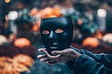 A pair of hands holds a black mask, the symbol of who society believes they are. The actually person stands out of frame. Afraid to expose themselves to the world without their mask.