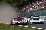 Dos coches de carreras Porsche compiten lado a lado en el circuito de Spa-Francorchamps. El coche de la izquierda es rojo y blanco con el número 6, mientras que el de la derecha es blanco con detalles en verde y el número 5. Detrás de ellos, otros vehículos siguen la acción de cerca. Al fondo, una multitud de espectadores observa la carrera, mientras una nube de polvo en el lado izquierdo indica la intensidad de la competencia.