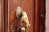 Wooden door with a bronze doorknob and a bronze hand sticking out to hold it.