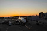 Commercial airplane parked at airport gate during sunset, connected to a jet bridge with ground crew and service vehicles nearby. Warm orange and yellow hues fill the sky, highlighting the serene yet active atmosphere of air travel operations.