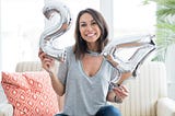 Brunette woman sitting on a white couch with silver “24” balloons for her birthday