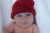 Baby (newborn) Stella with red knit hat, big blue eyes, yellow nasogastric tube smiling angelically at the camera