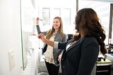 5 Ways to Increase Happiness in Your Employees image shows two women writing on a white board, smiling at one another.
