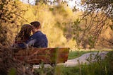 Two people are sitting on a bench on a summer or fall day. They’re smiling and their foreheads are touching.