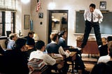 A quirky professor stands on two desks when speaking to students in English classroom