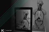 Person holding a tablet pointed at a medical training statue of a human with their internal organs expose. On the tablet, an augmented reality (AR) application is showing organ names in relation to the training statue. On the left side is the Kontor logo and a green Kontor watermark.