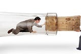 A man is trapped in the neck of a giant corked bottle laying on it’s side. Another man on the outside is pulling on the cork trying to open the bottle.