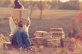 A lady reading next to a pile of books in the middle of a meadow.