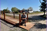 Don sits on a wooden bridge at the RVICS Village in Smithville, Texas, smiling on a sunny day with RVs and blue skies in the background.