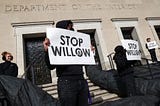 a person standing in all black at the department of the interior holding a sign that says “stop willow”