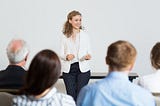 Mulher sorrindo para pessoas sentadas em sala de capacitação empreendedora