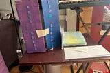Cardboard box painted purple and blue with two vertical lines of plastic bears glued to the front. A piece of tape is holding the box closed. A fuzzy notebook and a pile of envelopes are on the tray next to the box.