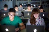 A photograph of nine students sitting in a dark classroom, all working on Apple laptops.