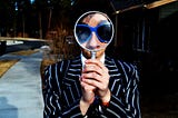 Man in pin-stripe suit and blue sunglasses looking through a large magnifying glass to promote SEO for About Now Ltd