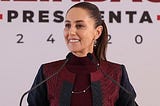 Mexican president Claudia Sheinbaum stands behind a podium with two microphones. Her dark hair is slicked back in a ponytail. She is wearing a maroon turtleneck with a maroon and black jacket, small earrings and a thin necklace.