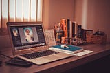 A laptop, a cellphone, a notepad, and some books set up on a wooden table in a home office.