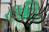 The mural features a mass of green paint splashed across the previously dilapidated wall behind a leafless tree, giving the appearance of the tree’s foliage. A person holding what seems to be a pressure hose has been stenciled alongside it. The artwork has just officially been confirmed as a Banksy; after many had already speculated as such as the mural resembled the style of the street-artist’s previous work.