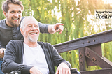 A photo of a younger man pushing an older gentleman in a wheelchair outdoors, both smiling and enjoying nature