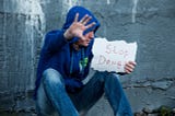 Young white man in a blue hoodie and jeans, sitting with knees up next to a gray concrete wall, holding his hand up with a sign that says, “Stop drugs”