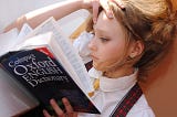 Photo of a light-skinned teenage girl with chestnut hair swept up in a loose bun, wearing long silver earrings, a white blouse, red tartan plaid suspenders and yellow polka dot tie, one hand on her head, reclined and reading a navy blue copy of the “Compact Oxford ENGLISH Dictionary”
