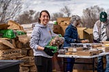 Color photo of food distribution volunteers preparing to share food with the community. A volunteer weraing a gray hoodie and wearing black gloives is holding a large sack of potatoes and smailing.