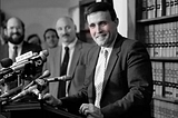 Black and white photo of Rudy Giuliani at a press conference in the 1987. Altered so he appears to be blushing.