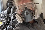 A very dirty person with blonde hair and pink skin is wearing a respirator and safety glasses while standing in front of a large pile of trash bags.