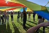 children and student volunteers playing with a rainbow parachute on a sunny day