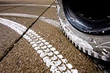 A pavement with a white paint imprint of a wheelchair tire tread. The powered wheel of the wheelchair is visible making the imprint.
