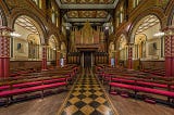 Image of the mid-Victorian chapel with lush red velvet seated wooden pew benches, gilded columns and pillared arches, a large gilded pipe organ at the very back and a pathway patterned tiles.