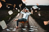 Barefoot man sitting on a chair in the middle of a country road. He’s covering his head with an open book from a “rain” of loose paper pages falling around him.