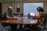 Monk (Jeffrey Wright) and Sintara (Issa Rae) having a discussion at a conference table.